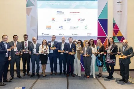 Group of professionals on stage holding trophies at an awards event, with sponsor logos on a large screen behind them.
