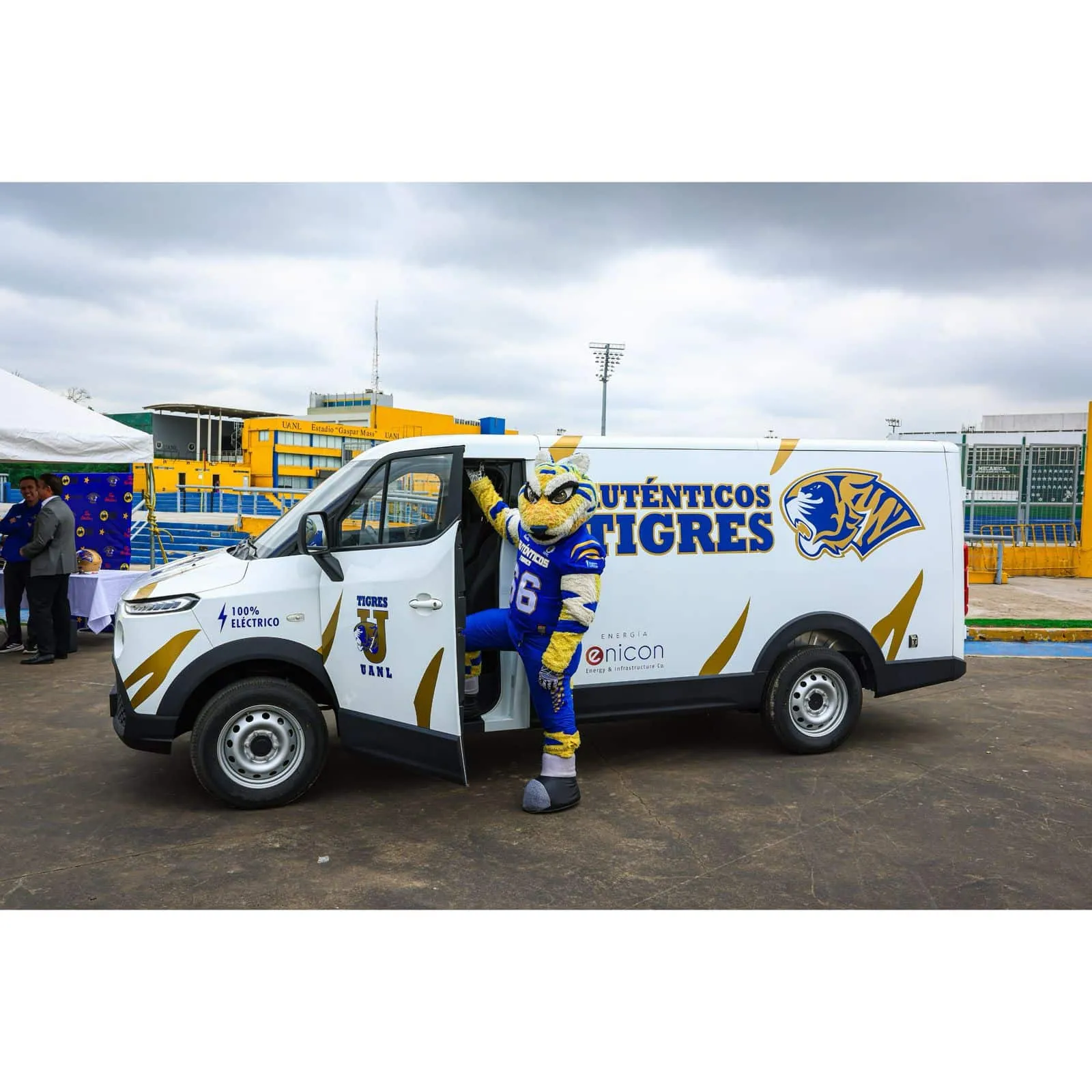 Mascot in blue and yellow uniform leaning out of a branded white van with Tigres logo at an outdoor event.