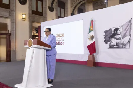 Woman in a lavender pantsuit speaks at a white podium on a stage, with a Mexican flag and a large screen reading '2026 año de Margarita Maza' in the background.