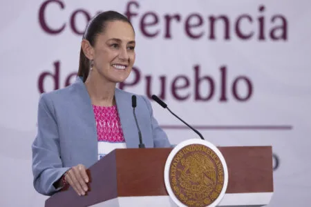 Female speaker at a podium with the Mexican seal during a conference, smiling as she addresses the audience.