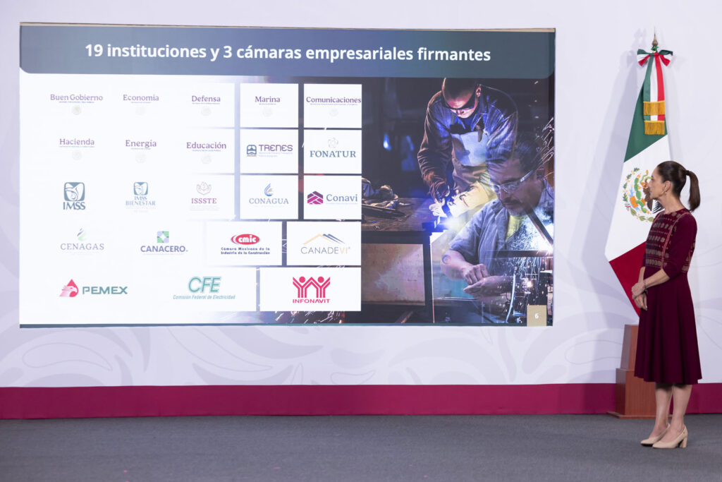 2026-04-29_Conferencia_de_prensa_matutina-Palacio_Nacional_04_GM - Petróleo y Energía Mujer con vestido rojo de pie junto a un cartel grande con logos de 19 instituciones y 3 cámaras empresariales firmantes; bandera mexicana a la derecha.