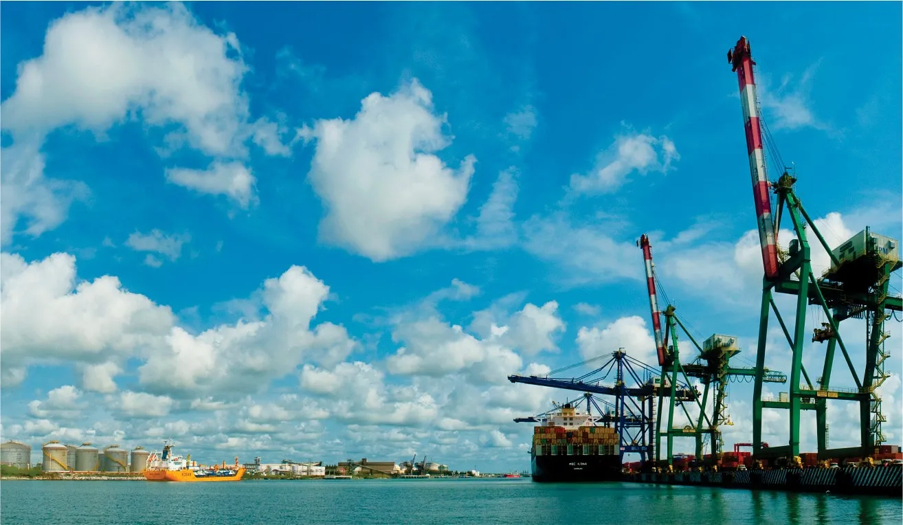 Container ship docked at a busy port with large green gantry cranes loading containers under a bright blue sky with clouds.