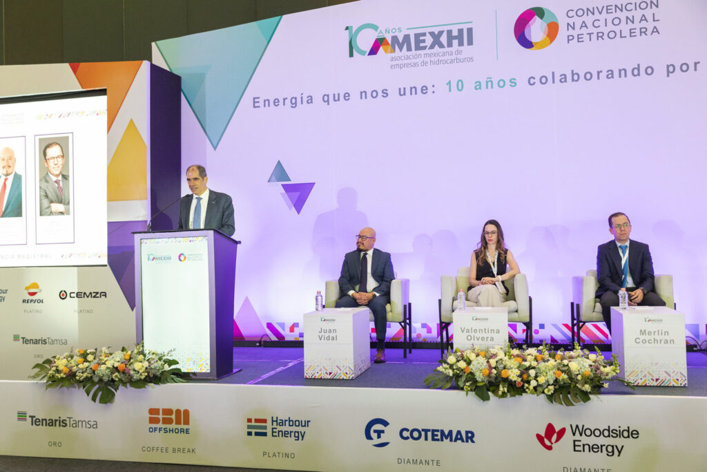 54452815110_f452c1ee4e_k-1536x1024 - Petróleo y Energía Man in suit speaks at a lectern on a colorful conference stage, with three panelists seated and floral arrangements in front.