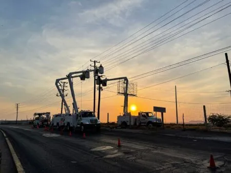 Utility crews with bucket trucks working on power lines at sunset along a cracked road, traffic cones lining the street.
