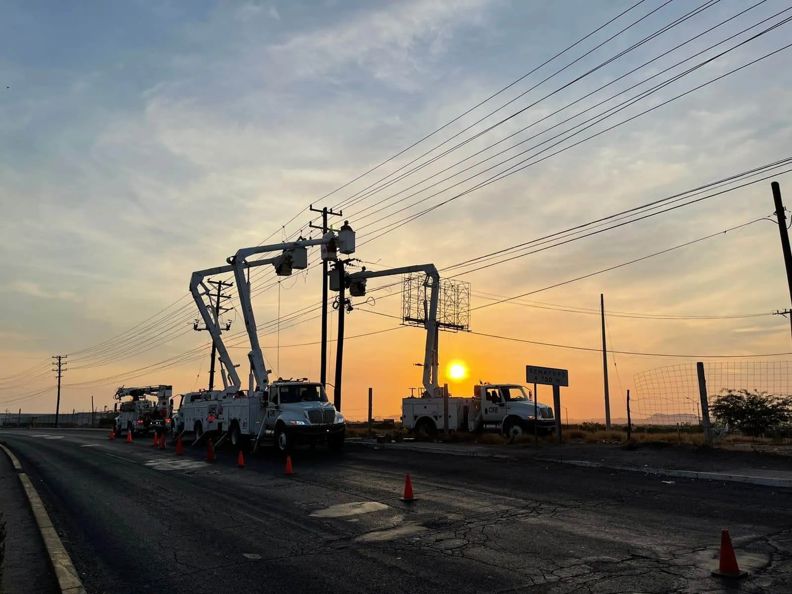 Utility crews with bucket trucks working on power lines at sunset along a cracked road, traffic cones lining the street.