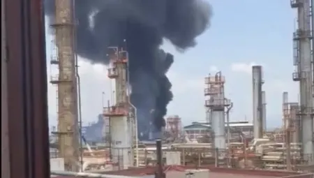 Industrial refinery with a dark smoke plume rising from a central stack amid nearby towers and structures.