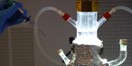 Scientist wearing blue gloves injects a syringe into a lab reactor with tubing and foil-wrapped insulation, under bright light.