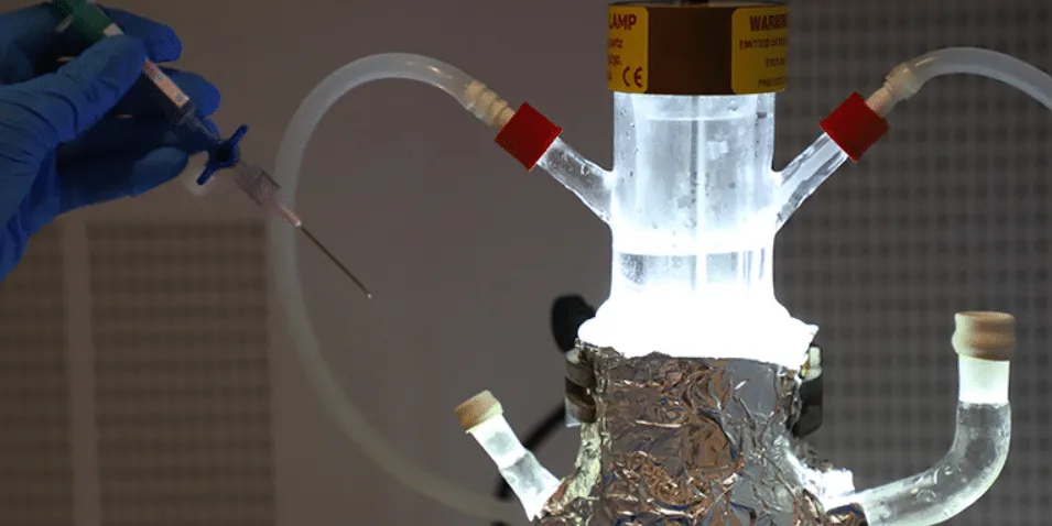 Scientist wearing blue gloves injects a syringe into a lab reactor with tubing and foil-wrapped insulation, under bright light.