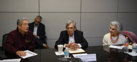 Panel of three adults seated at a conference table in discussion; center man in suit speaks, others listen with papers and bottles nearby.