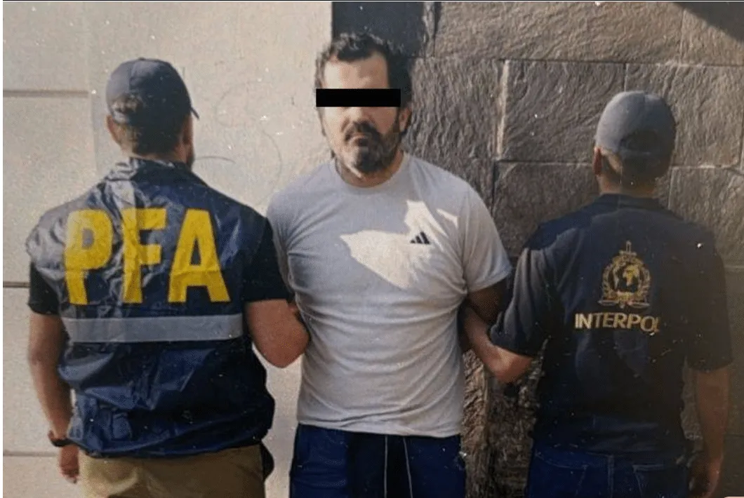 Man with a black bar over his eyes is escorted by two officers, one wearing a PFA jacket on the left and the other an Interpol jacket on the right, from behind and side view.