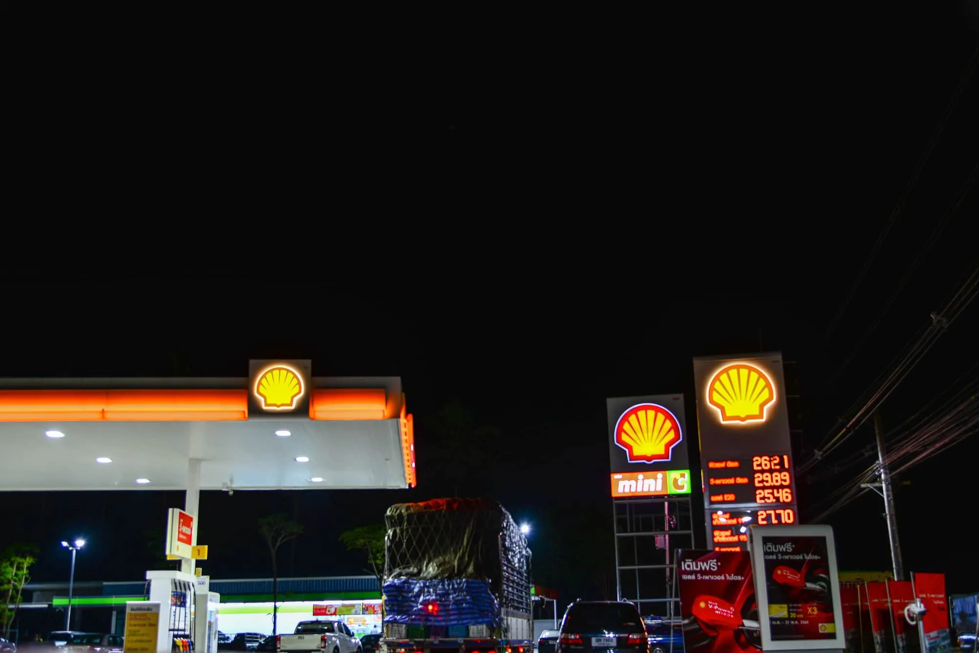 Nighttime Shell gas station with bright orange signs and logo, pumps visible and cars parked nearby.