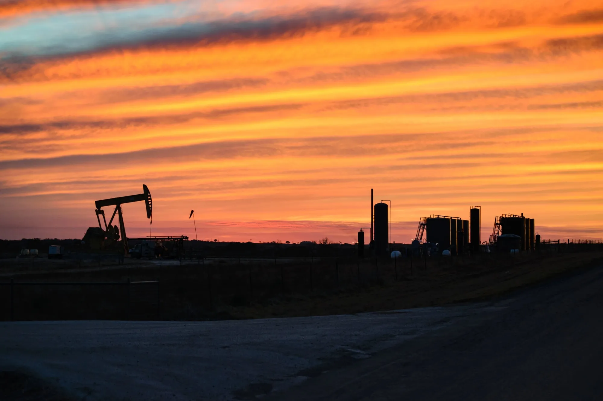 Oil pumpjack silhouette at sunset with orange sky and industrial buildings on the horizon