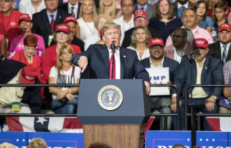 Male politician in a dark suit and red tie speaks at a podium with the presidential seal in front of a cheering crowd wearing red hats.
