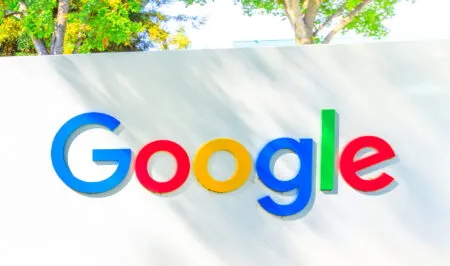 Google logo on a light wall with colorful letters and trees in the background (outdoor sign).