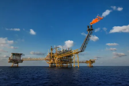 Offshore oil platform in the sea with a tall flare stack burning bright orange flames under a blue sky.