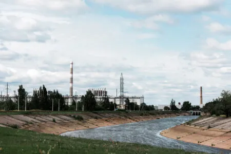 Industrial landscape with tall smokestacks and factory buildings along a waterway under a cloudy sky, trees in the foreground.