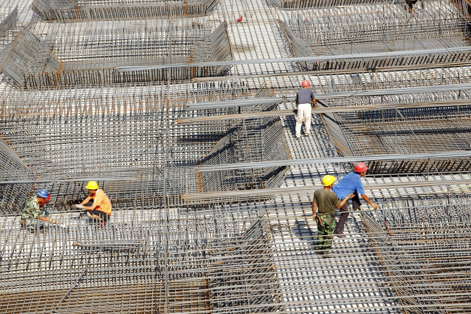 Construction workers in hard hats walk on a dense grid of steel reinforcement bars at a building under construction.