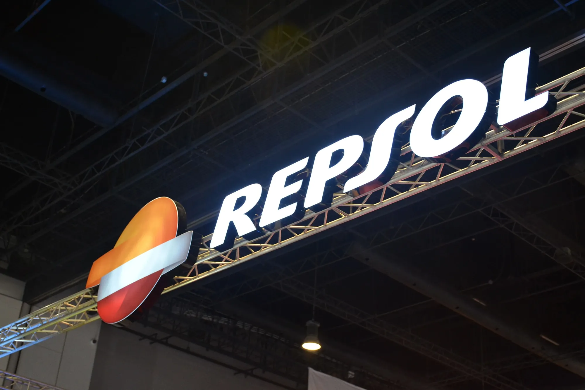 Repsol brand sign on a metal truss at an expo, with the white 'REPSOL' text beside an orange-red circular emblem with a white stripe, illuminated.