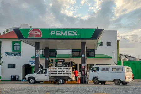 Pemex gas station forecourt with a green canopy sign, fueling pumps, and vehicles, including a white van and a small truck in the foreground.