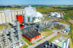 Aerial view of a large industrial plant with silos, tanks, and a plume of steam rising from a chimney, set against fields in the distance.