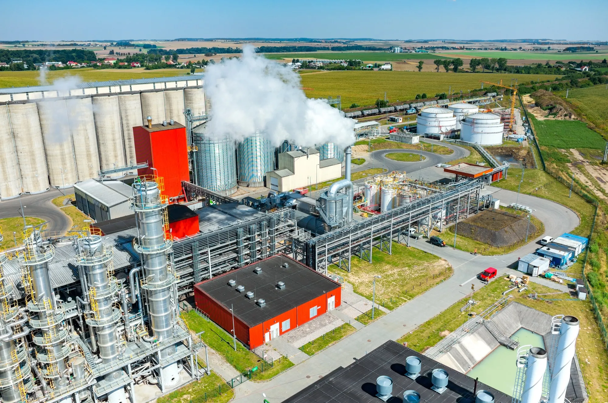Aerial view of a large industrial plant with silos, tanks, and a plume of steam rising from a chimney, set against fields in the distance.