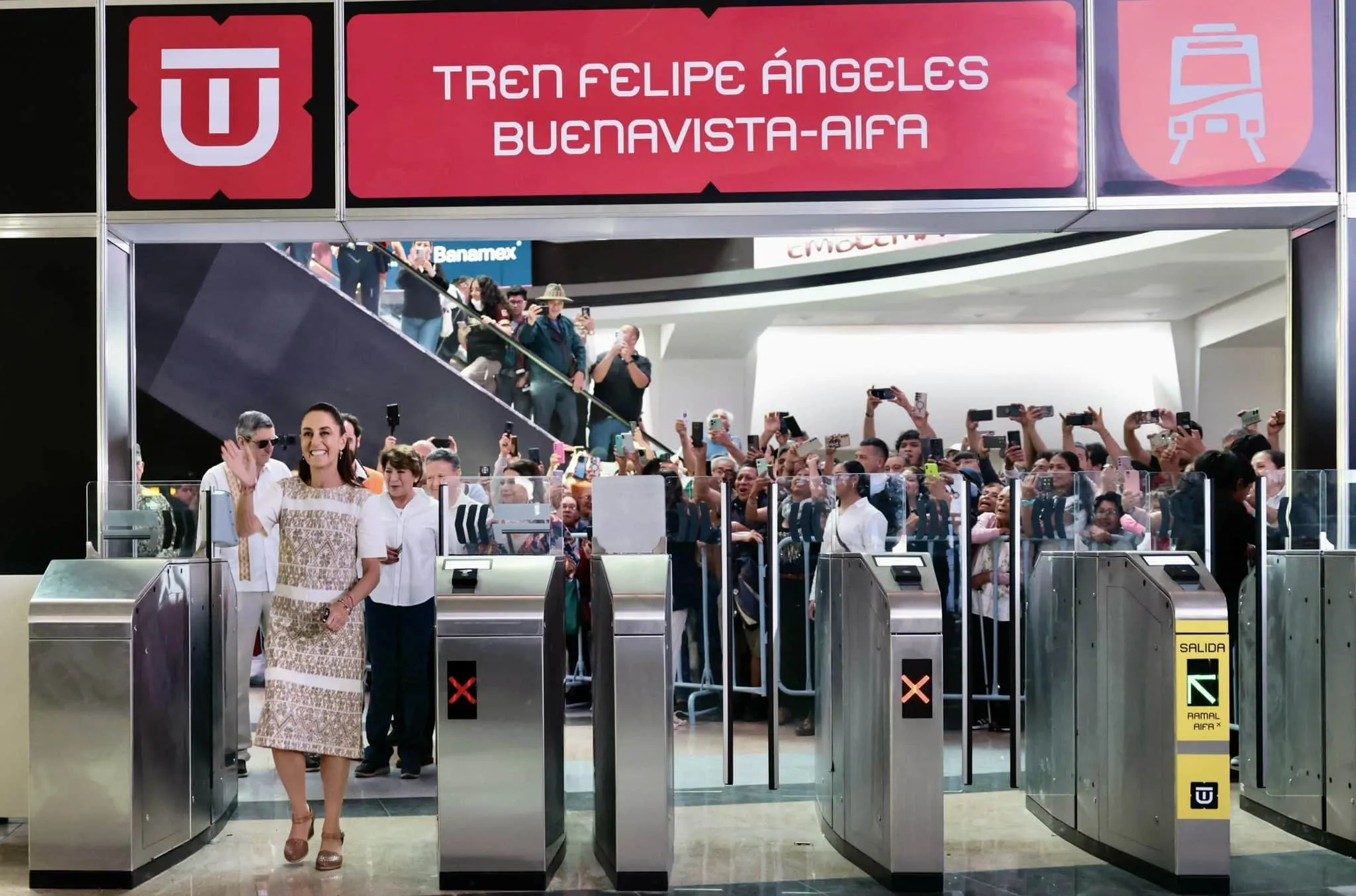 Smiling woman in a patterned dress stands at metal turnstiles in a crowded transit terminal with a pink banner above.