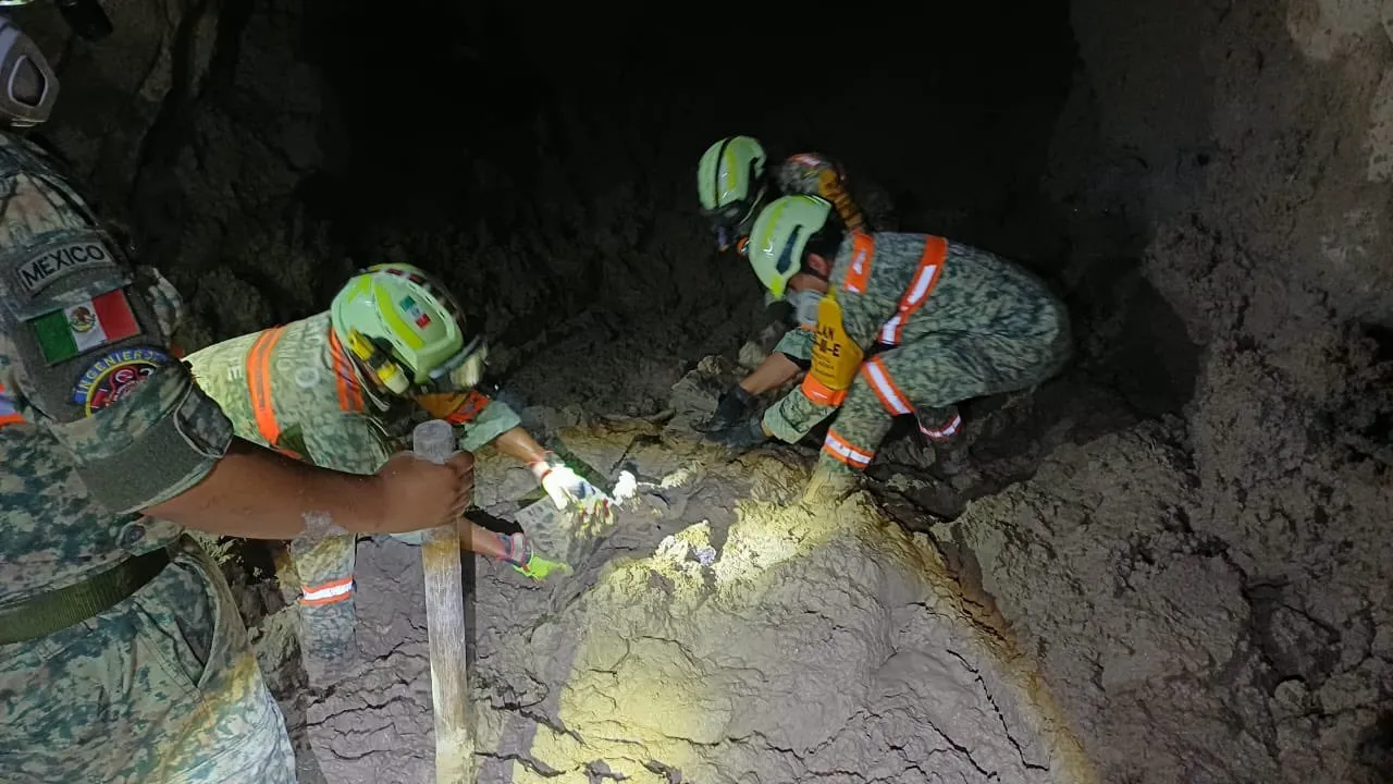 Three Mexican disaster-response rescuers in helmets and reflective vests work inside a dark, muddy cave, using tools to probe a crack in the rock while a headlamp illuminates the area.