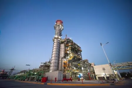 Industrial refinery complex with a tall processing tower and interconnected pipes at dusk, lights on.
