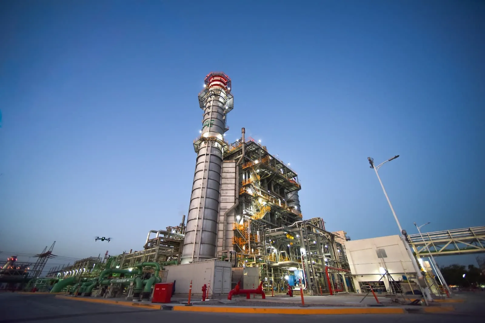 Industrial refinery complex with a tall processing tower and interconnected pipes at dusk, lights on.