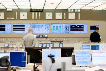 Control room with multiple large monitors showing data dashboards and charts, and two technicians at the back monitoring systems.