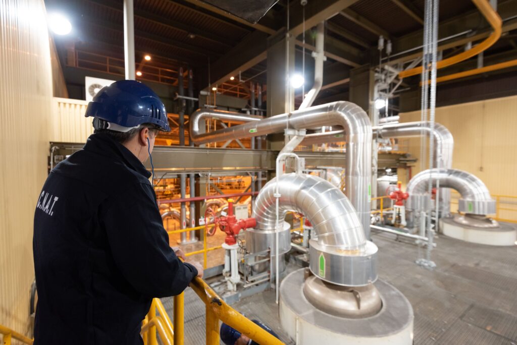 HGWCTzcWcAEcaEO - Petróleo y Energía Worker in a blue hard hat and dark protective jacket stands on a yellow railing, overlooking large silver industrial pipes and valves in a factory setting