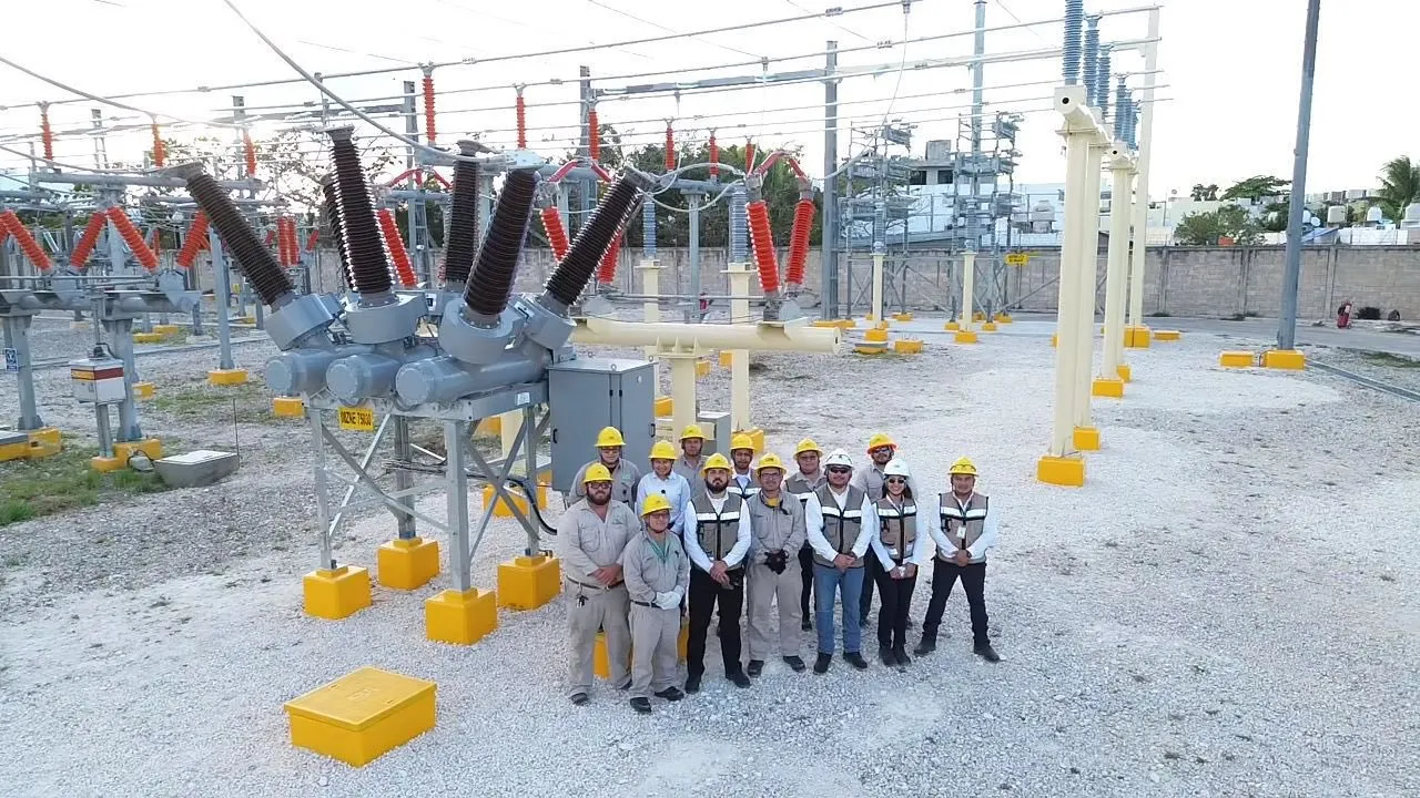 Group of workers in hard hats and safety vests standing together at a power substation site with large electrical equipment behind them, posing for a photo.