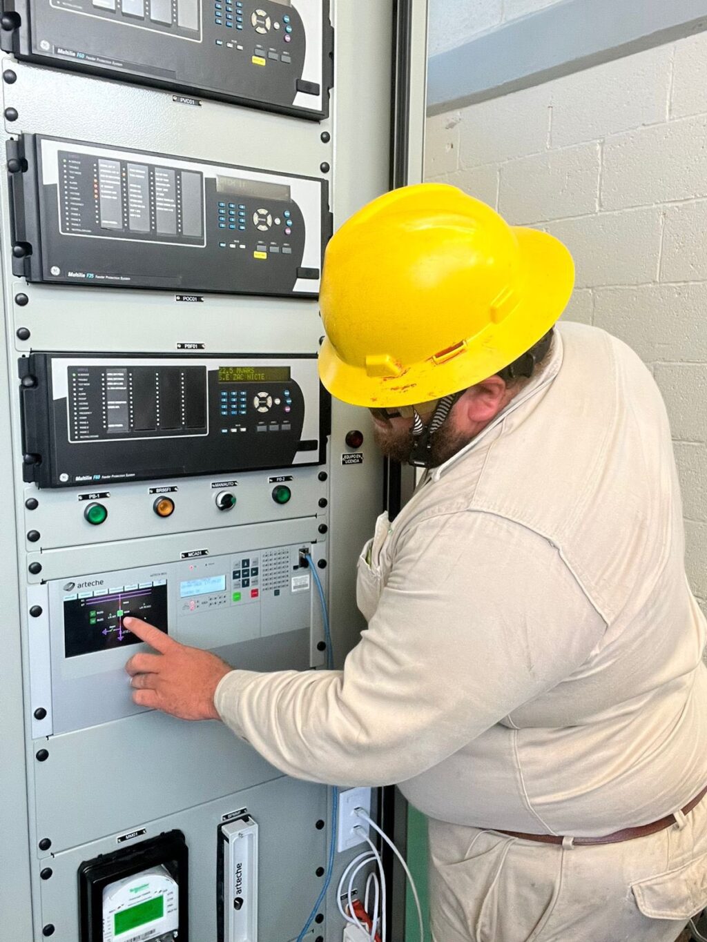 HGXf13tasAASJFa - Petróleo y Energía Maintenance worker in a yellow hard hat operating a tall electrical control cabinet with display screens and buttons in a utility room.