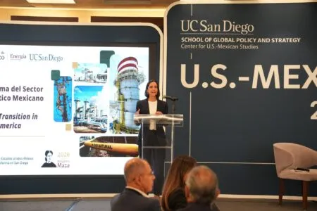 Speaker at a podium during a conference with UC San Diego branding and a large U.S.-Mexico banner in the background.