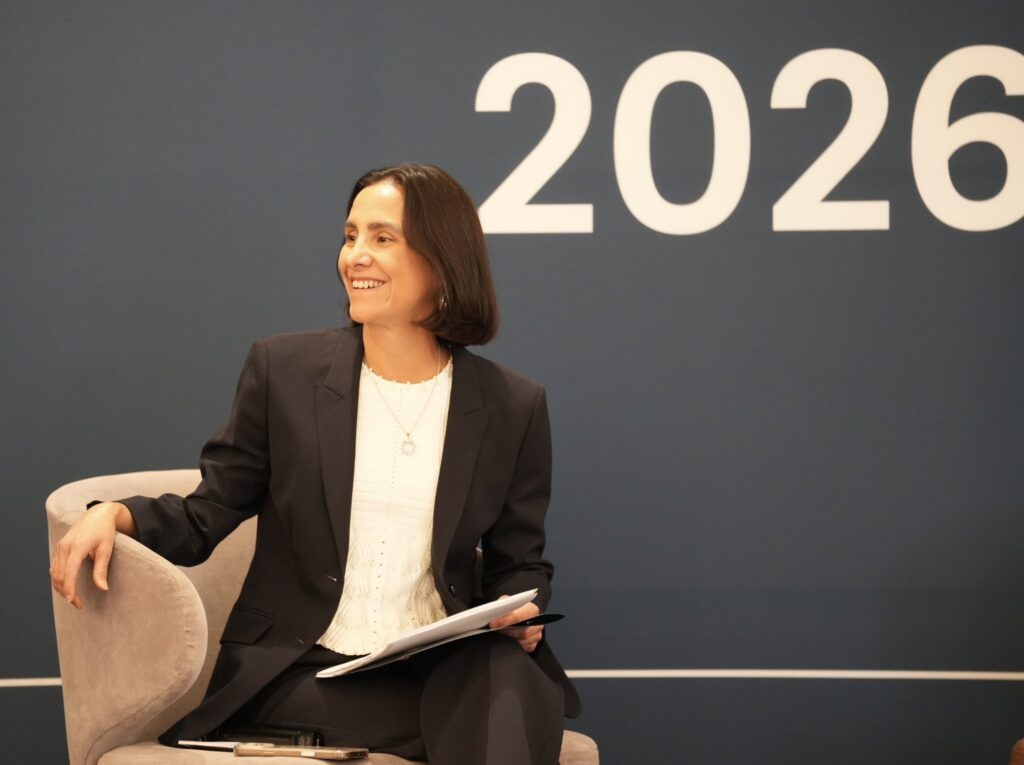 HGYhqdQaAAAl6rR - Petróleo y Energía Smiling woman in a dark blazer seated on a beige chair, holding papers, with a large 2026 backdrop behind her on stage.