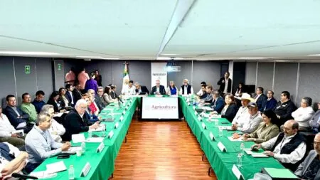 Large conference room with dozens of people seated around U‑shaped green tables for an agricultural meeting; screen reads 'Agricultura'.