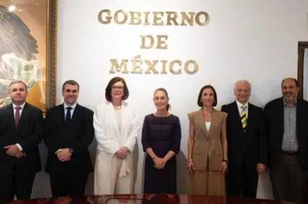 Eight formally dressed individuals standing side by side for a group photo in front of a wall displaying gold letters that read 'GOBIERNO DE MEXICO'.