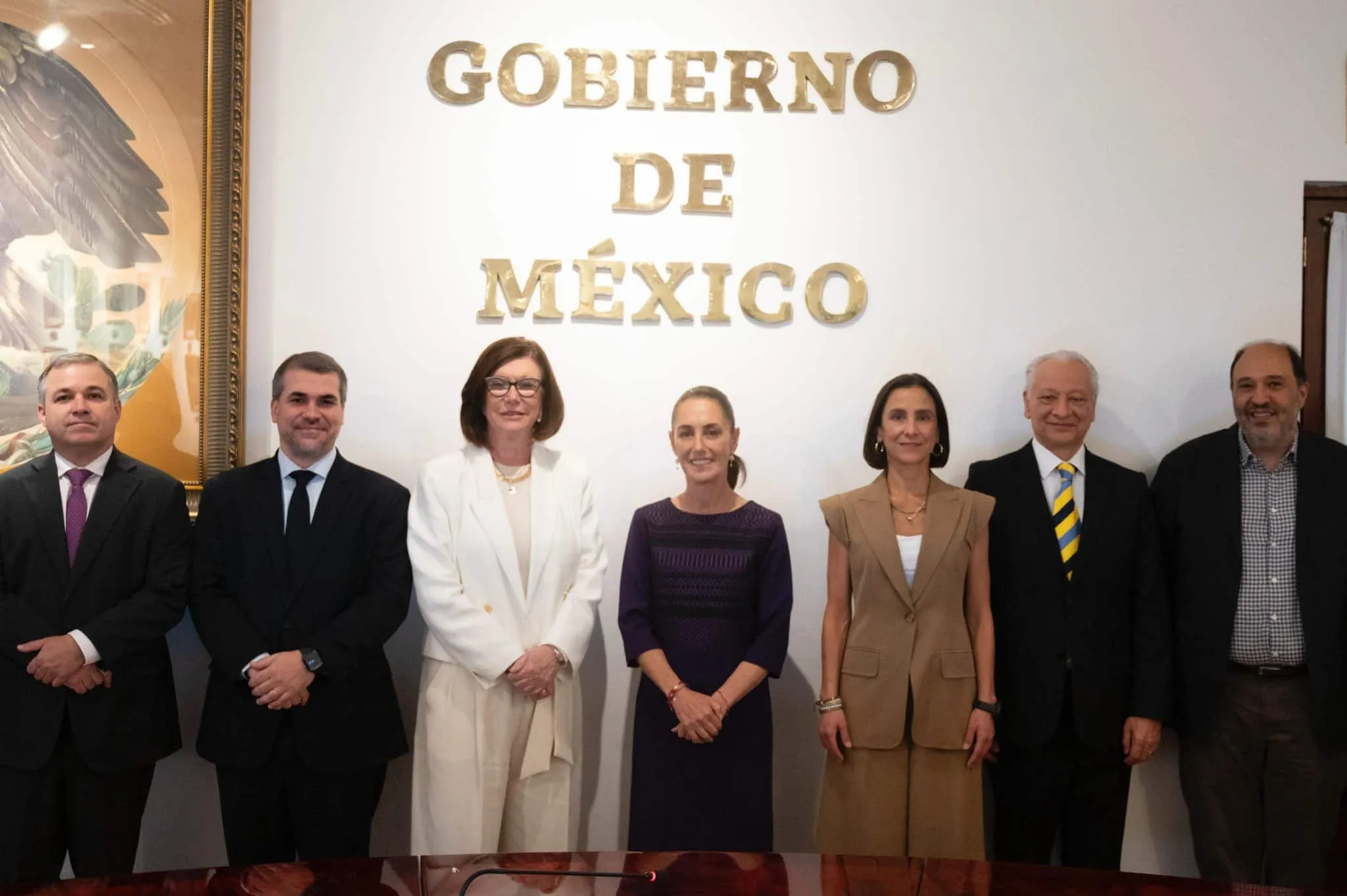 Eight formally dressed individuals standing side by side for a group photo in front of a wall displaying gold letters that read 'GOBIERNO DE MEXICO'.