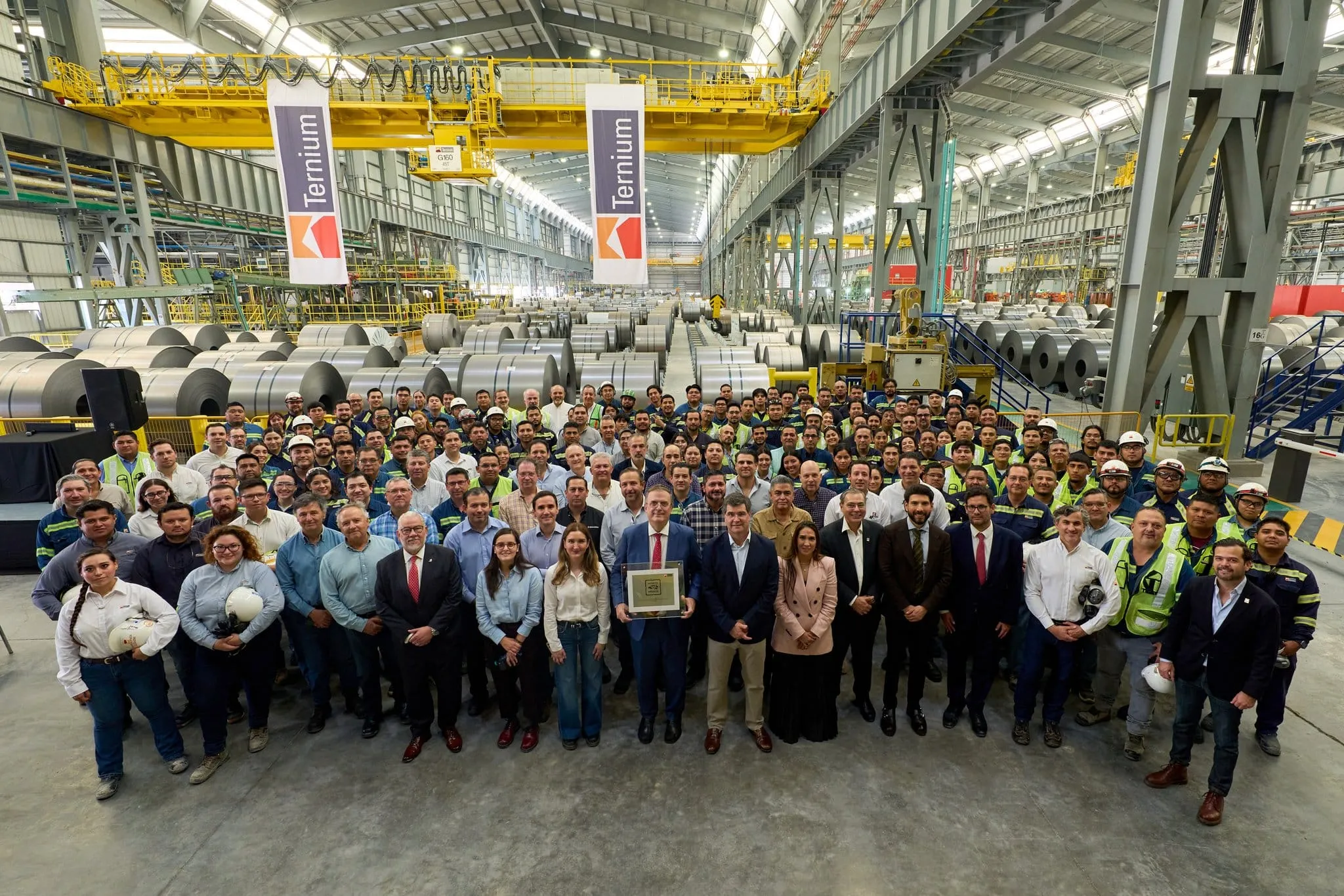 Group of factory workers and managers posing for a photo inside a large steelmaking/fabrication plant with coiled metal rolls and yellow overhead cranes in the background.