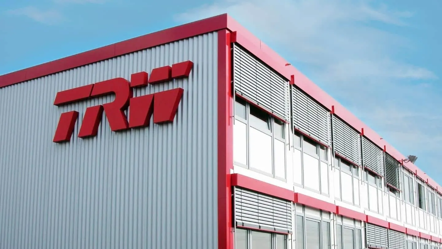 Industrial building with a bold red TRW logo on gray corrugated metal siding and red trim under a blue sky.
