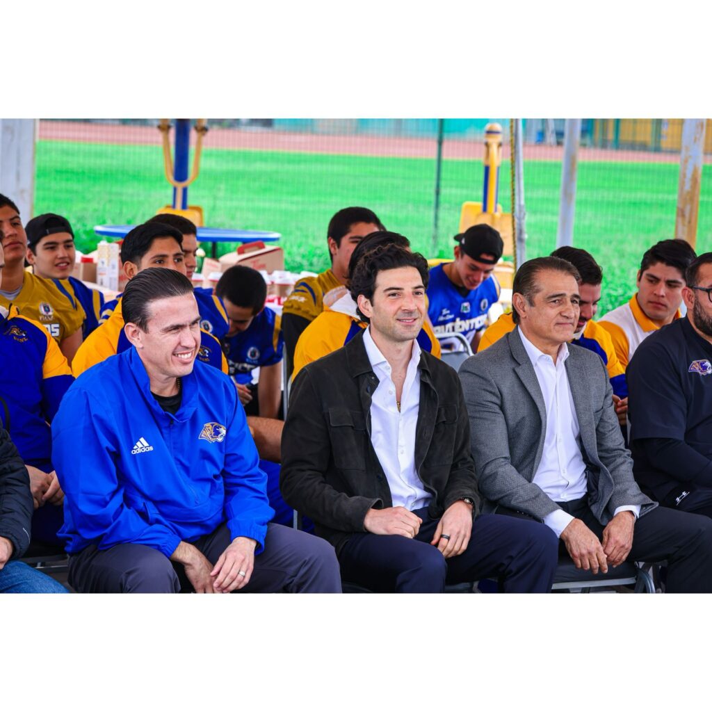 fb8796f0-3c9c-4195-9208-8e3d4dbad4a2 - Petróleo y Energía Group of men seated in a row at an outdoor event; some wear blue/yellow team jerseys, others in suits, with a green field in the background