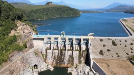 Concrete dam with multiple spillways releasing water into a rocky chute, across a blue reservoir surrounded by hills and distant mountains.