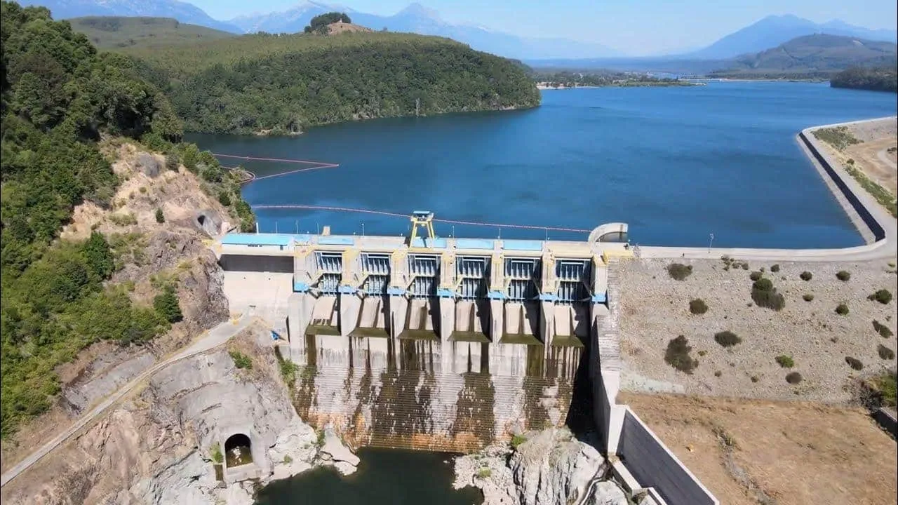 Concrete dam with multiple spillways releasing water into a rocky chute, across a blue reservoir surrounded by hills and distant mountains.