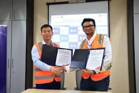 Two men in orange safety vests shake hands and hold signed documents in a conference room.