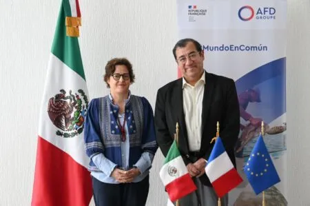 Two officials stand side by side smiling in front of Mexican, French, and EU flags with a banner in the background.