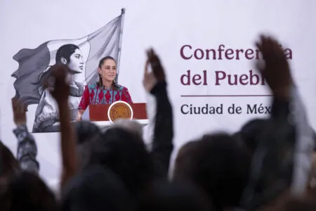 Woman at a podium giving a conference, with a large banner reading 'Conferencia del Pueblo' and a Mexican seal on the lectern behind her