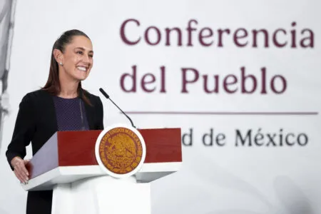 Woman speaking at a podium with the Mexican seal during a conference, smiling at the audience.