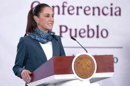 Woman in a teal blazer speaking at a podium with the Mexican seal, microphones in front, conference backdrop behind.