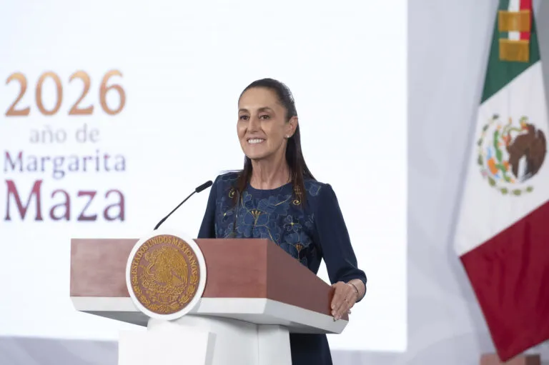 Female speaker at a wooden podium delivering a formal address, Mexican flag visible to the right and a seal on the front of the podium.