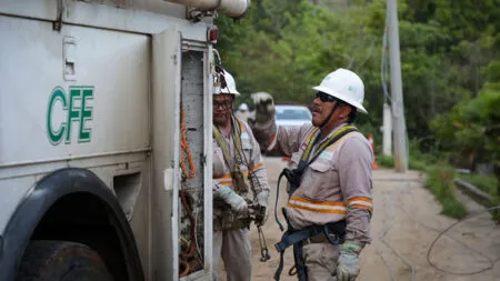 Two utility workers in hard hats and safety gear stand by a service truck marked CFE on a rural dirt road, talking.